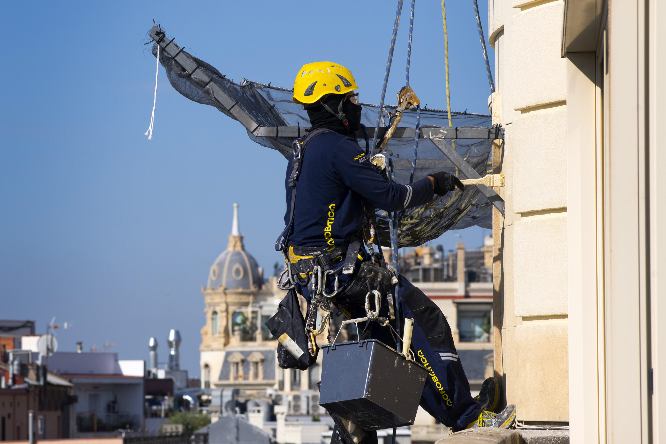 Operatore su fune con casco giallo maneggia una rete di protezione, con i tetti di Barcellona sullo sfondo